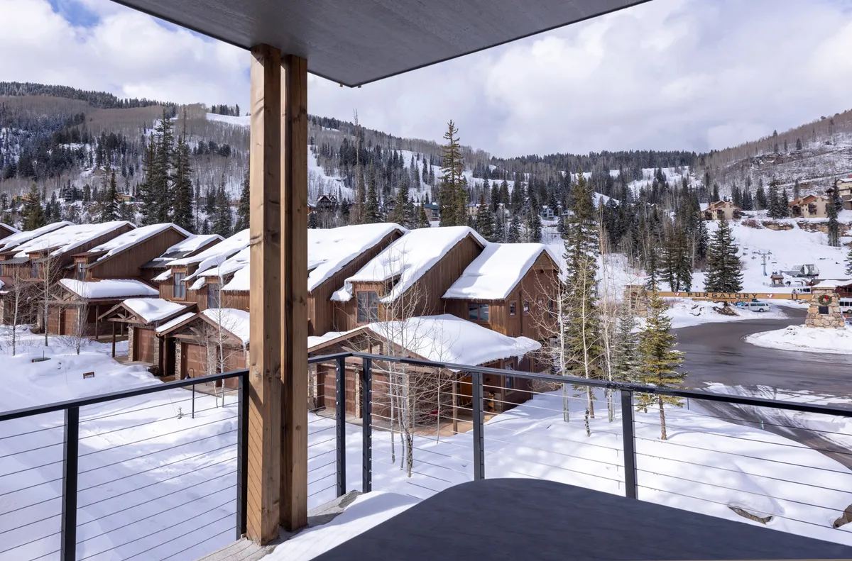 A serene view of snow-covered rooftops nestled among pine trees, all set before a dramatic mountain backdrop. The scene evokes feelings of coziness and adventure, hinting at relaxing mornings and exciting days spent outdoors.