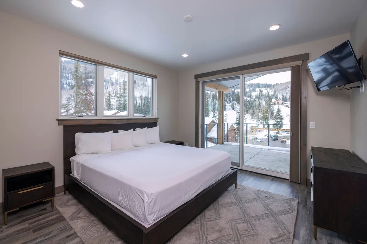 This interior image features a spacious bedroom with a comfortable bed, large windows, a door leading to a private balcony, and a TV. Soft natural light fills the room, creating a tranquil sanctuary perfect for rest and relaxation after a day of adventure.