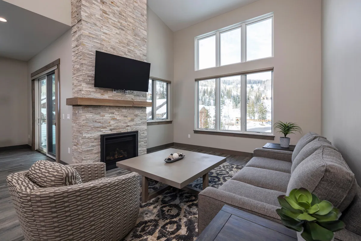 The image showcases a stylish, modern living room with a striking stone fireplace as the centerpiece. Expansive windows fill the space with natural light and offer a glimpse of the scenic outdoors. A plush, inviting couch and contemporary coffee table create a cozy atmosphere perfect for relaxing or spending time with family and friends.