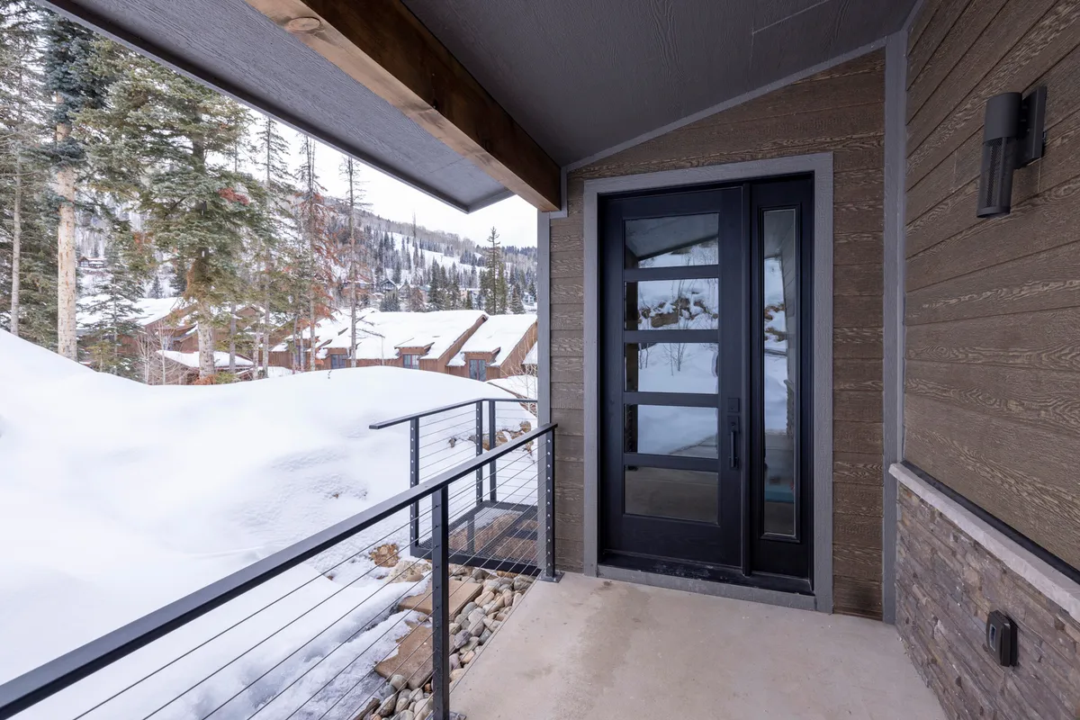 This image captures the front door of the property framed by a tranquil, snow-covered landscape, with railings leading up to the entry and mountain views in the distance. The inviting scene hints at both a warm welcome and immediate access to the outdoors.