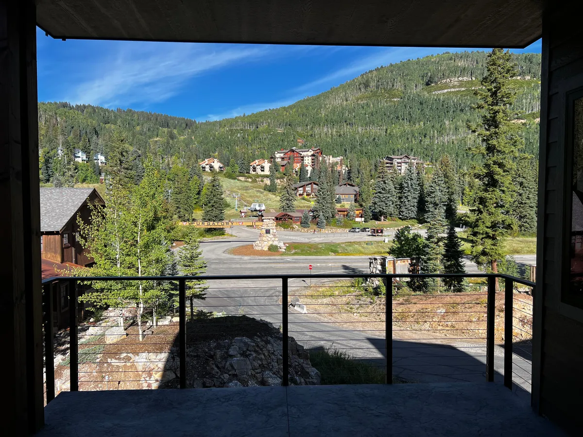 A sweeping view of a mountain landscape with colorful buildings nestled at the base, bordered by a lush tree line. A road leads toward the mountains, suggesting easy access to hiking, nature exploration, and community activities.