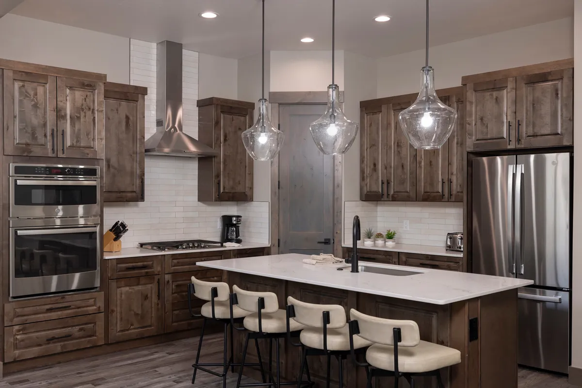 A bright, stylish kitchen featuring a spacious island at the center. Stainless steel appliances and wooden cabinetry line the walls, with elegant hanging light fixtures above creating a warm, inviting glow. The open layout connects seamlessly to other living areas, hinting at a sociable and flexible space perfect for cooking and entertaining.