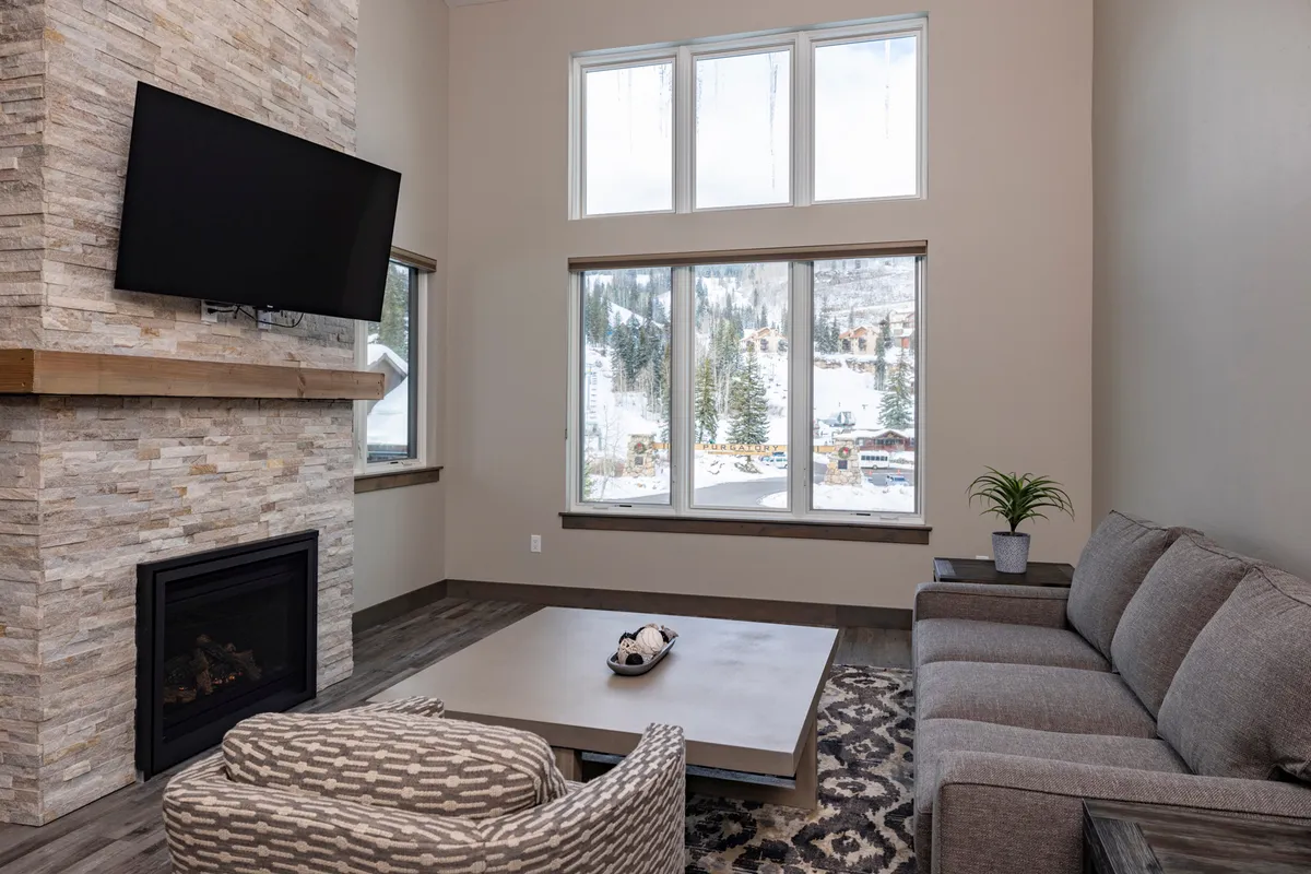 A cozy living room featuring a plush sofa arranged around a stone fireplace, with expansive windows showcasing a snowy mountain landscape. The coffee table and comfortable seating invite relaxation and conversation, while the warm glow of the fire creates a welcoming après-ski atmosphere.