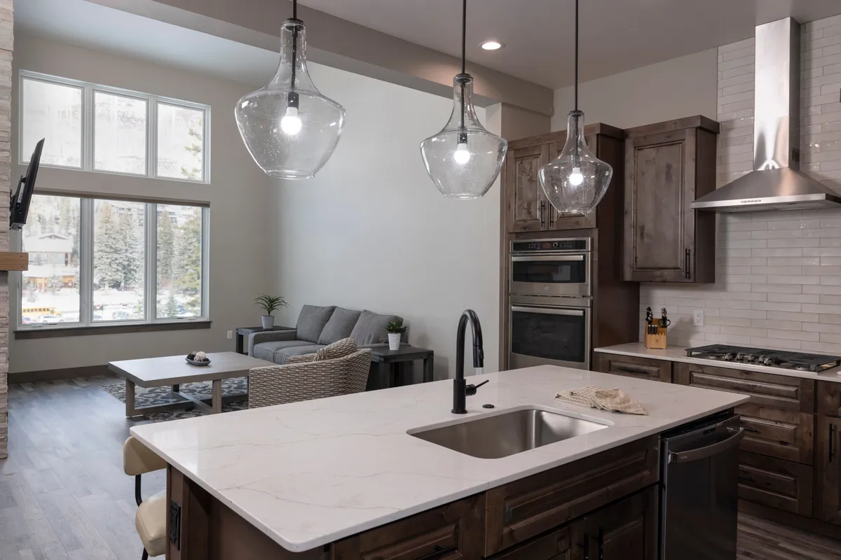 A stylish modern interior combining a comfortable seating area, a kitchen island with pendant lights, and expansive windows that flood the space with light and offer views of the outdoors. The open plan ensures easy movement and socializing, with direct lines of sight to the scenery outside.