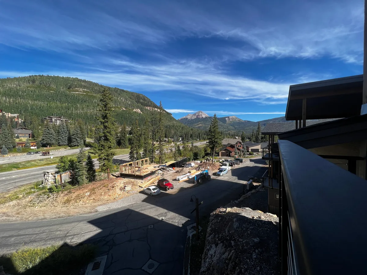 This image captures a sweeping view of the mountain range, with a tree line, a roadway, parked cars, and hints of nearby construction—framing the stunning natural scenery and the vibrant activity of the local community.