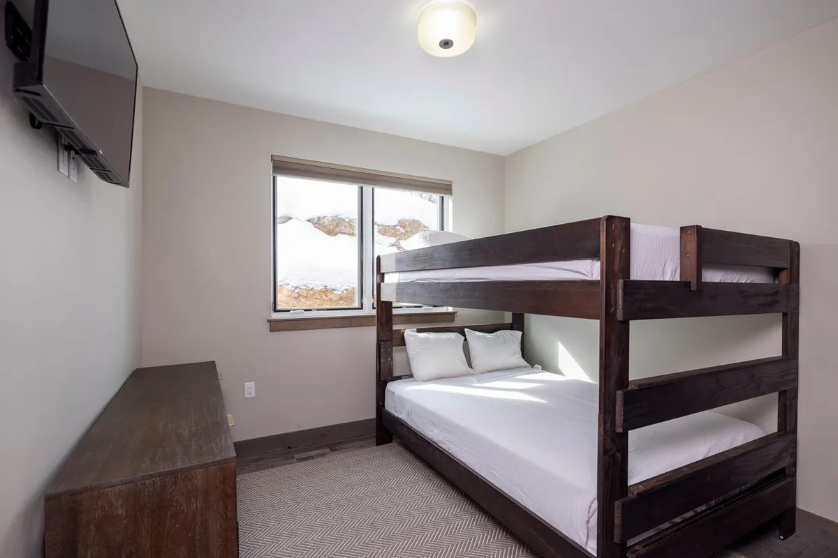 A cozy bunk room featuring a queen-sized bunk bed, a window for natural light, a television, and a dresser. The room offers a playful yet comfortable setting ideal for families or groups traveling with children.