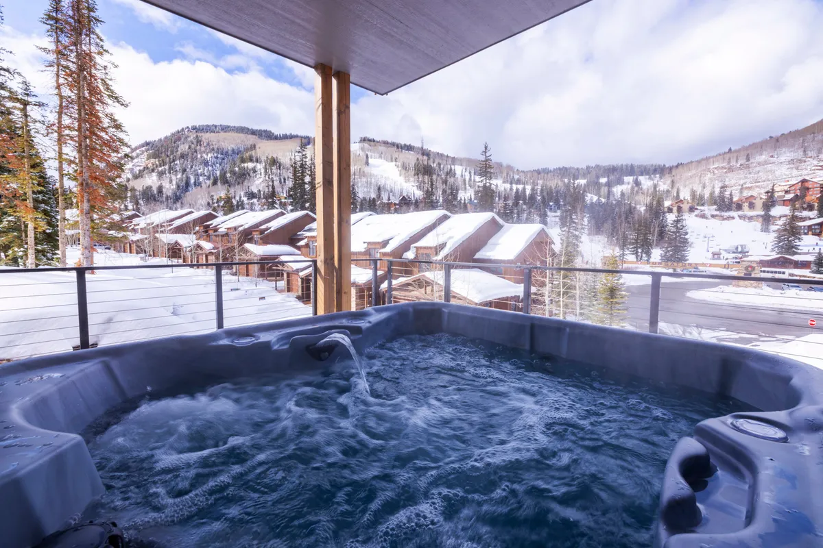 The image features an inviting outdoor hot tub set against a stunning backdrop of snow-covered mountains and wooden cabins. The ambiance is tranquil, with the promise of relaxation while soaking in the heated water and gazing at the expansive mountain scenery.