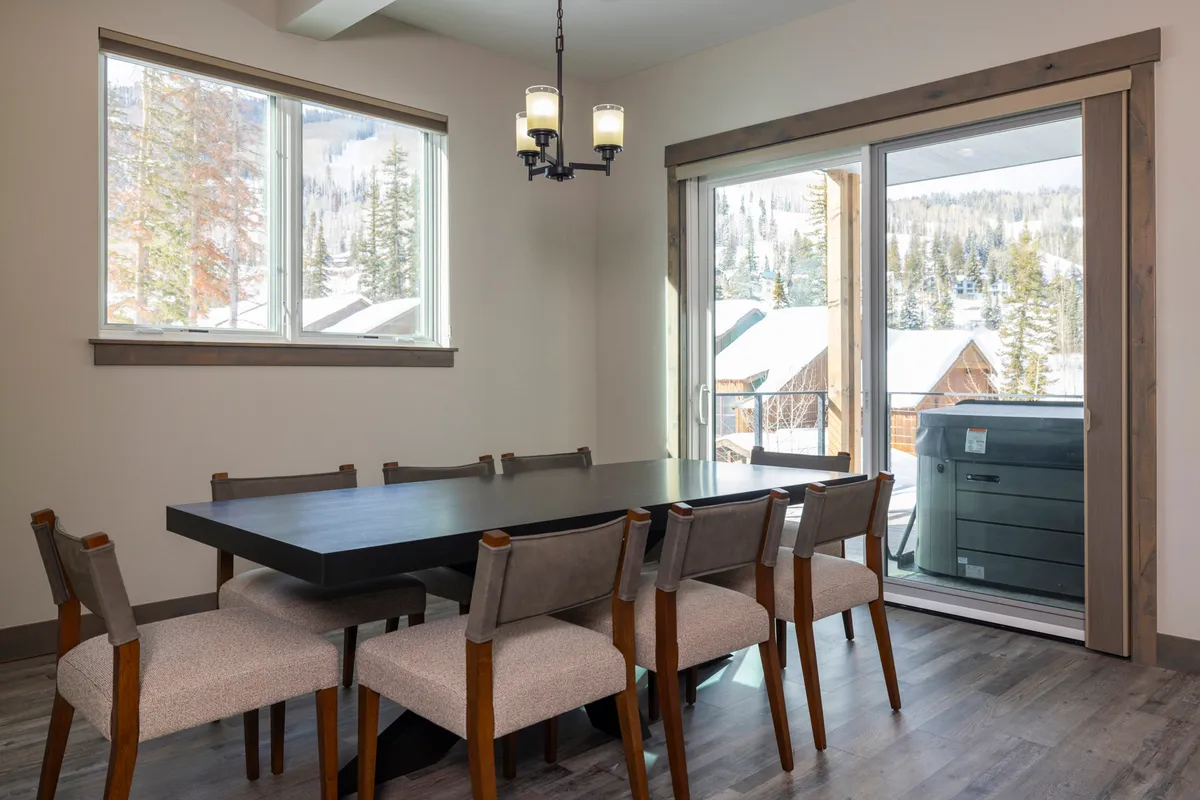 A dining area with a large table set beneath a glowing chandelier, positioned in front of floor-to-ceiling windows that offer clear views of the snowy landscape outside. The inviting space is perfect for group meals, celebrations, or game nights, with natural light enhancing the cozy mood.