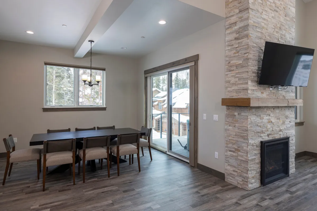 A cozy dining area with a large table set near a crackling fireplace. Sliding glass doors in the background bring in natural light and possibly offer glimpses of scenic views. The space radiates warmth and togetherness, making it ideal for gathering with friends or family over meals and conversation.